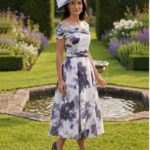 Elegant woman in a blue‑gray floral off‑the‑shoulder midi dress, matching hat, and purple heels walking in a formal garden with a fountain in the background.