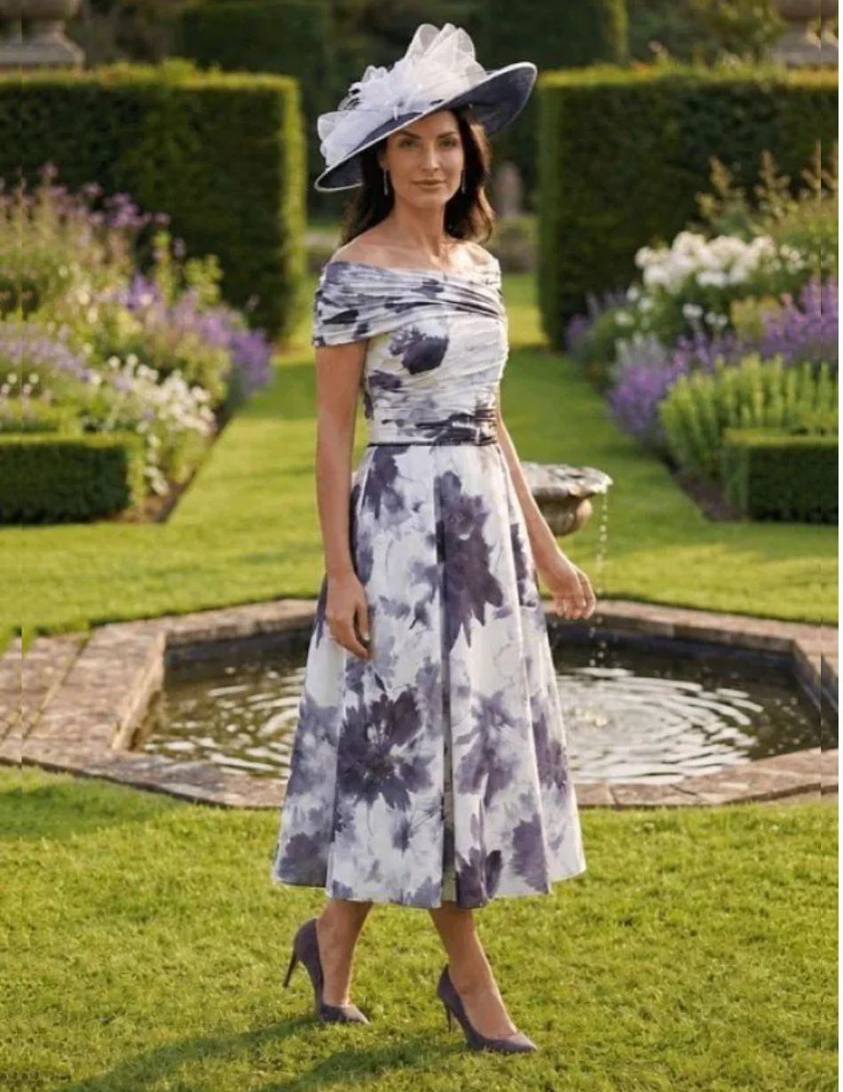 Veromia VO3144 Navy Elegant woman in a blue‑gray floral off‑the‑shoulder midi dress, matching hat, and purple heels walking in a formal garden with a fountain in the background.