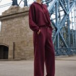 Woman in a burgundy matching hoodie and wide-leg pants stands with hands in pockets beside a blue steel bridge, looking into the distance.