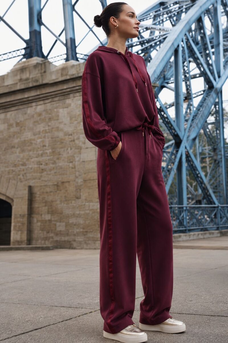 Woman in a burgundy matching hoodie and wide-leg pants stands with hands in pockets beside a blue steel bridge, looking into the distance.