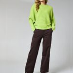 Woman modeling a bright lime-green knit sweater with dark brown wide-leg pants and black boots, standing against a light backdrop.